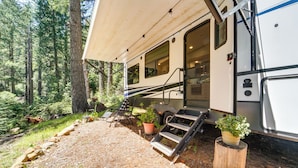 A recreational vehicle parked in a forested area with a covered porch, outdoor seating, and potted plants. - Wooded Getaway w/ Beautiful Views in Nevada City! (Nevada City)