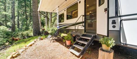 A recreational vehicle parked in a forested area with a covered porch, outdoor seating, and potted plants.