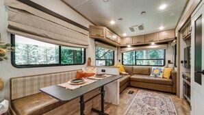 A compact RV interior with a kitchenette, seating area, and large windows offering views of the outdoors. - Wooded Getaway w/ Beautiful Views in Nevada City! (Nevada City)