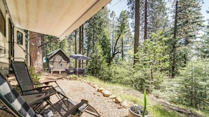A covered outdoor seating area with a chair and a table, a wooden cabin in the background, and a forested landscape. - Wooded Getaway w/ Beautiful Views in Nevada City! (Nevada City)