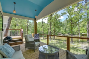 A covered outdoor deck with wicker furniture, a glass table, and a view of a wooded area.