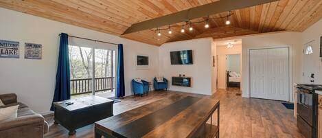 A spacious room with a wooden ceiling, a long dining table, a sofa, a TV, and a view of the outdoors.