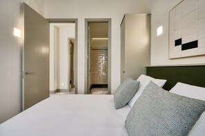 Ein modernes Hotelzimmer mit einem großen Bett, zwei Kissen, einem Kopfteil, einem an der Wand befestigten Kunstwerk und einem sichtbaren Duschbereich.