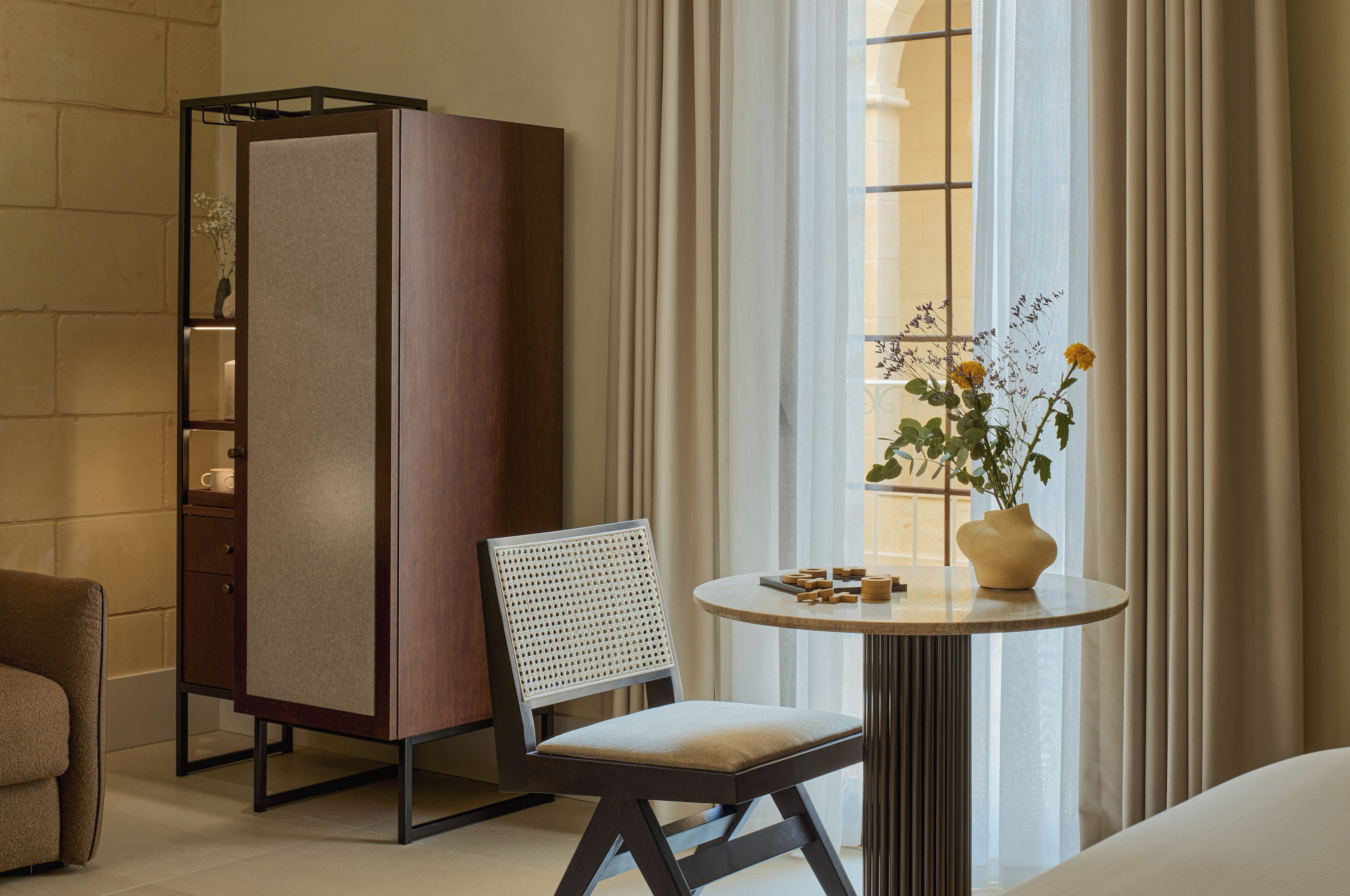 A hotel room with a bed, a seating area with a chair and a table, a wardrobe, and a window with curtains.
