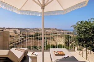 Un balcón con vistas a un paisaje montañoso y al mar. Hay dos sillones de descanso y una mesa con una bandeja de comida y bebidas.