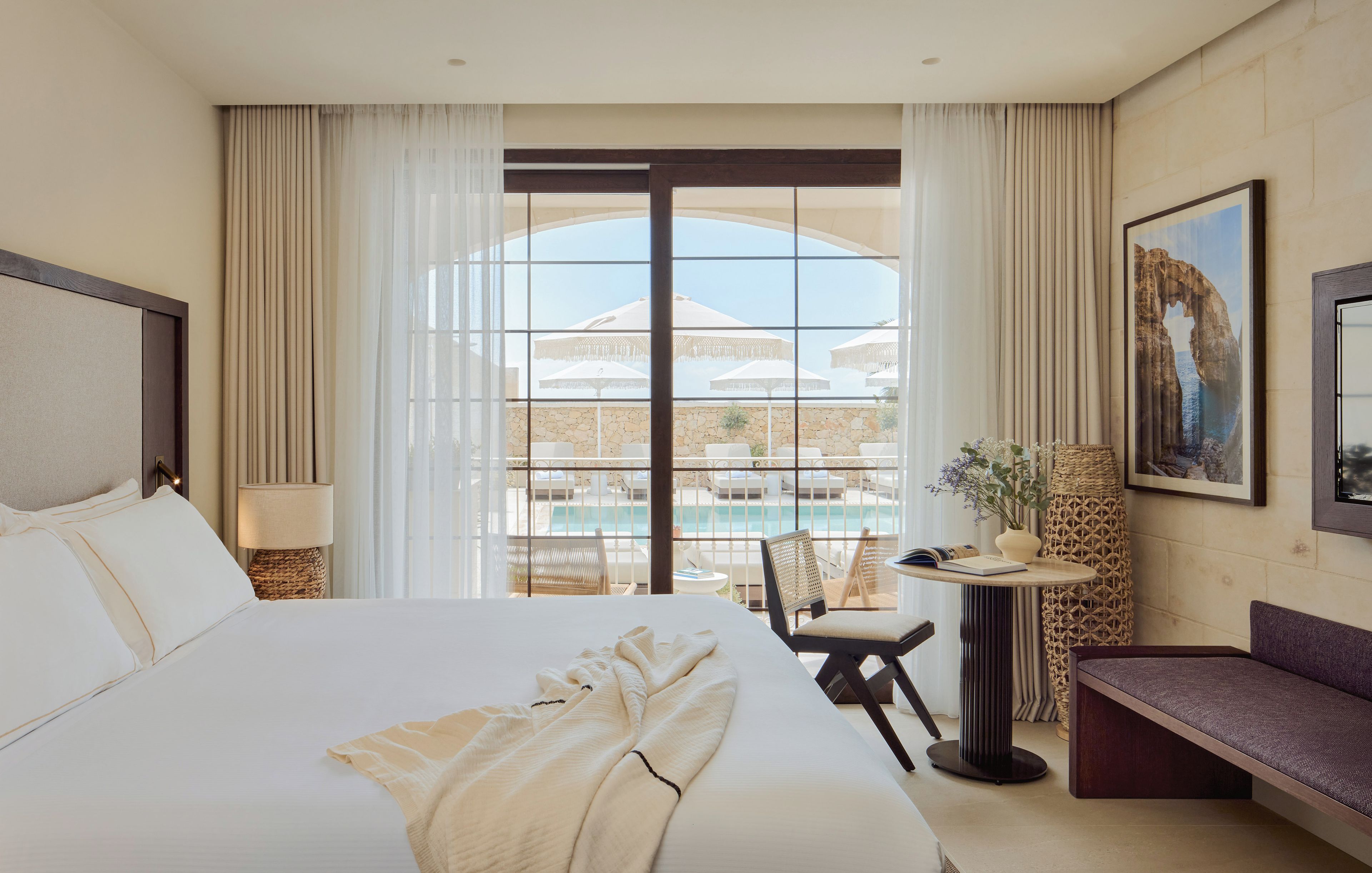 Habitación de hotel con una cama grande, un escritorio, una silla y vistas a una piscina y un edificio a través de la ventana.