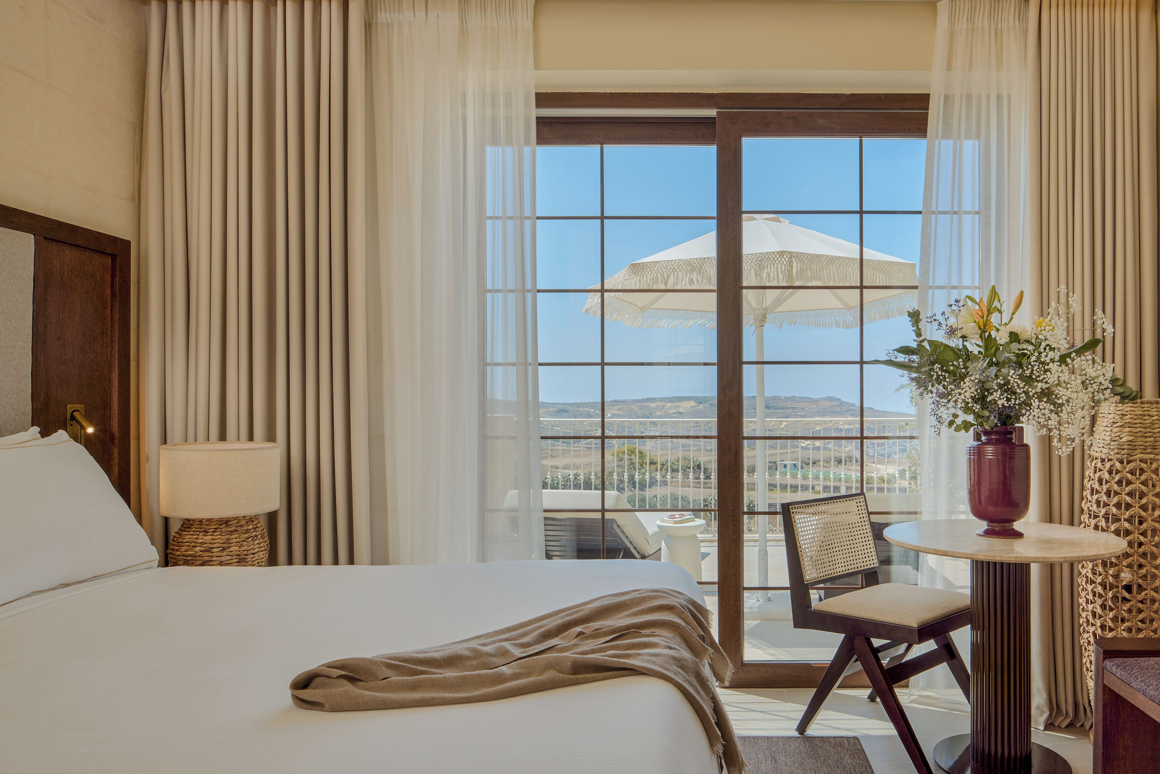 A hotel room with a bed, a table with a vase of flowers, a chair, and a balcony with an umbrella.
