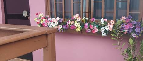 Un edificio con fachada rosa, una puerta de madera con un letrero con el número 5, una ventana de diseño calado y un arreglo floral en el alféizar.