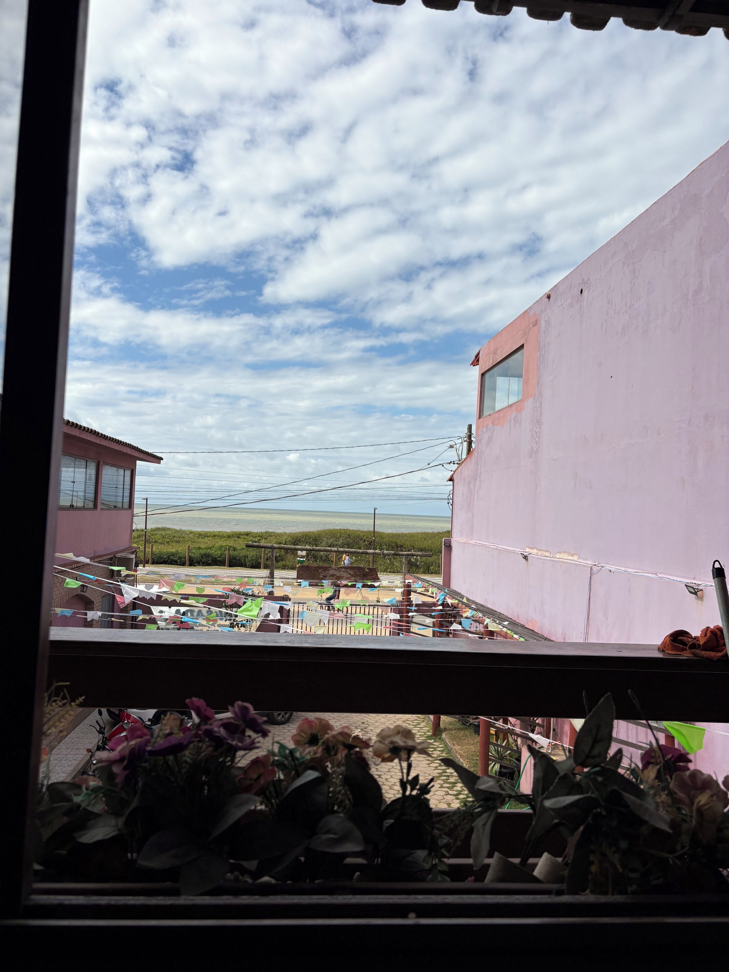 A view through a window of a building with a pink exterior, a balcony with plants, and a clear sky.