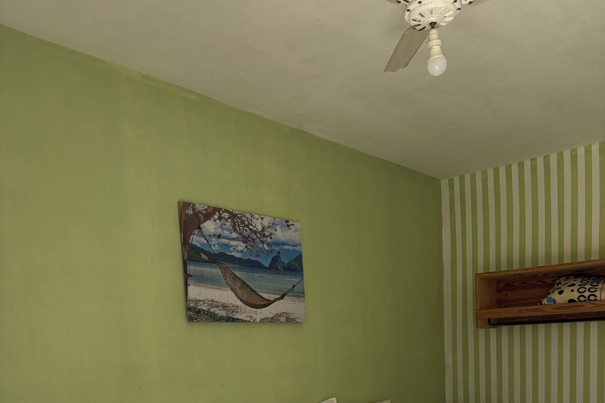 Habitación de hotel con dos camas, un ventilador de techo, un cuadro de una playa y una pared a rayas.