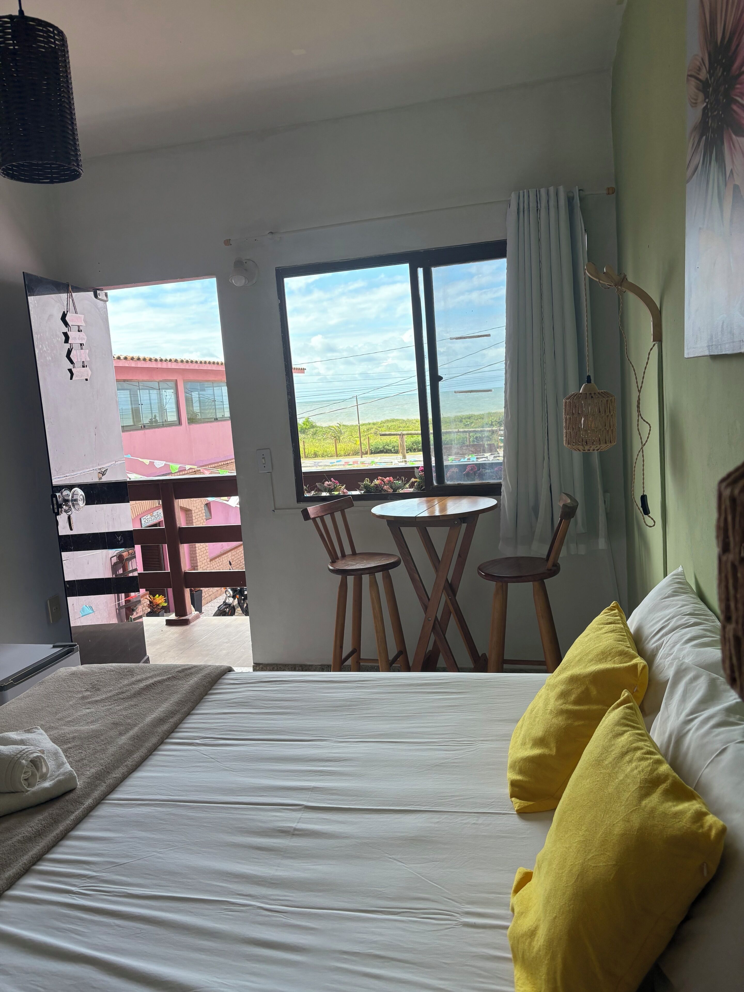 Habitación de hotel con cama, una mesita, dos sillas, ventana con vistas a edificios y una lámpara colgante.