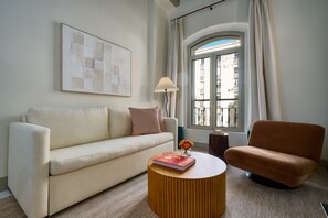 Un salon avec un canapé, une table basse ronde et un fauteuil. Il y a une fenêtre avec des rideaux, une lampe et un tableau encadré au mur.