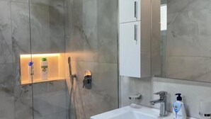 A modern bathroom with a glass-enclosed shower, a white sink with a mirror above, and a cabinet with a built-in shelf. - Villa Duka (Kruje)