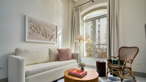 Ett vardagsrum med en soffa, ett runt soffbord, en fåtölj i rotting, en golvlampa och ett stort fönster med gardiner.