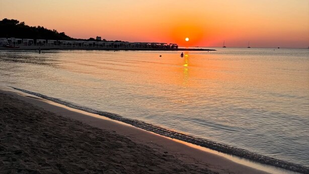 Een strand met een zonsondergang, rustig water en een rij gebouwen langs de kust.