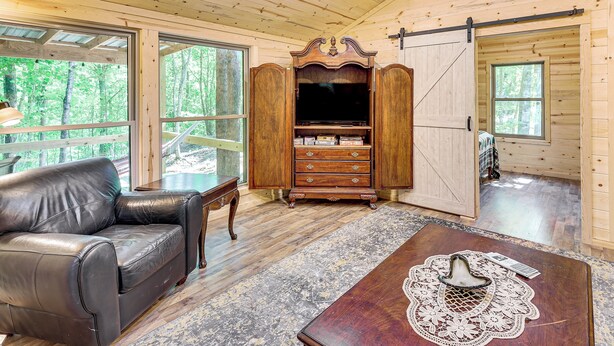 A wooden cabin interior with a leather sofa, a wooden entertainment unit, and a large sliding door. - Gateway to Ouachita National Forest! Cozy Cabin (Glenwood)