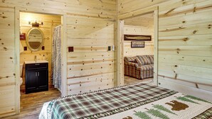 A wooden cabin interior with a bed, a bathroom with a sink and toilet, and a plaid-patterned rug. - Gateway to Ouachita National Forest! Cozy Cabin (Glenwood)