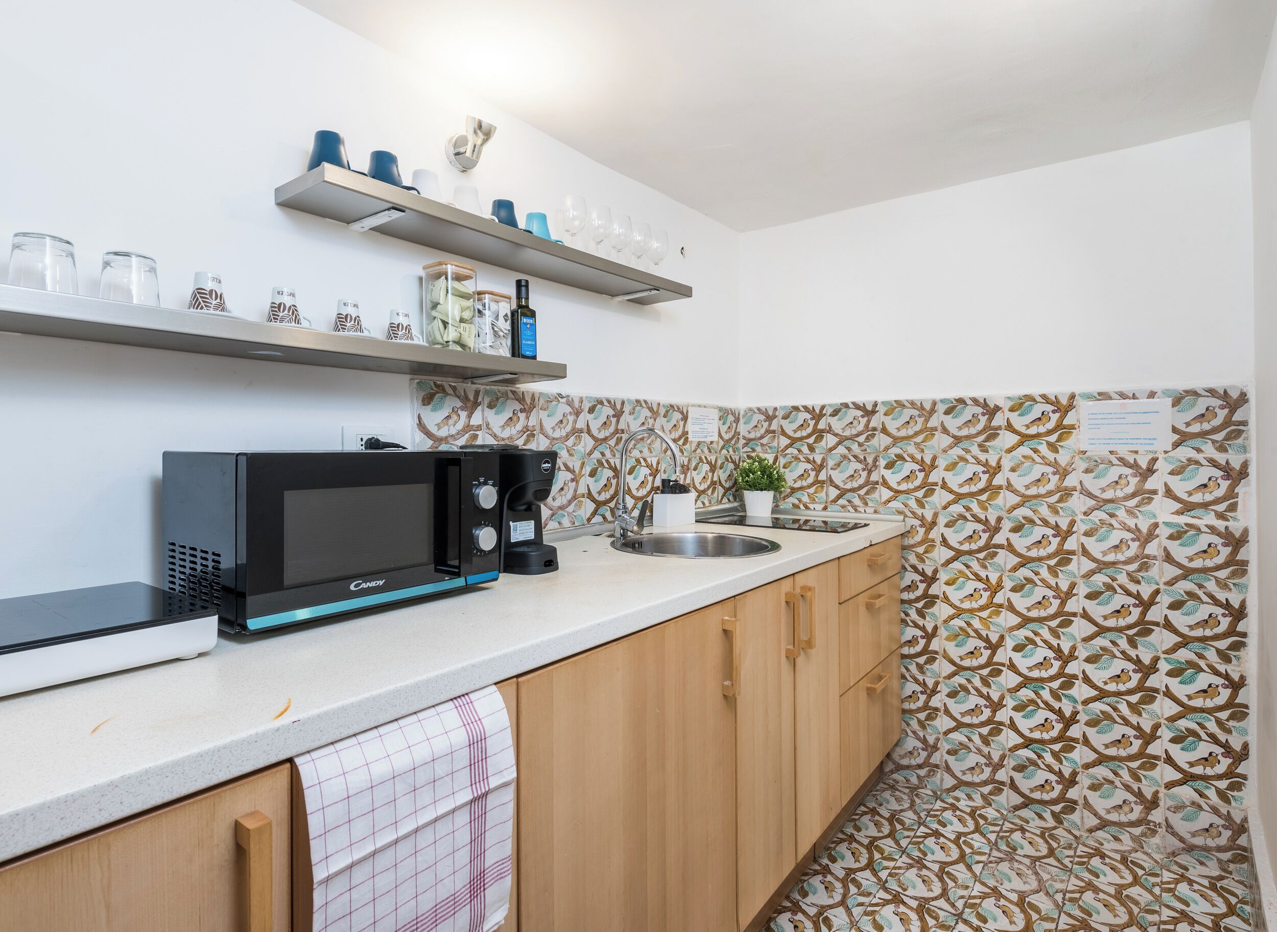 A kitchen with a microwave, coffee maker, and a shelf with glasses and bottles.