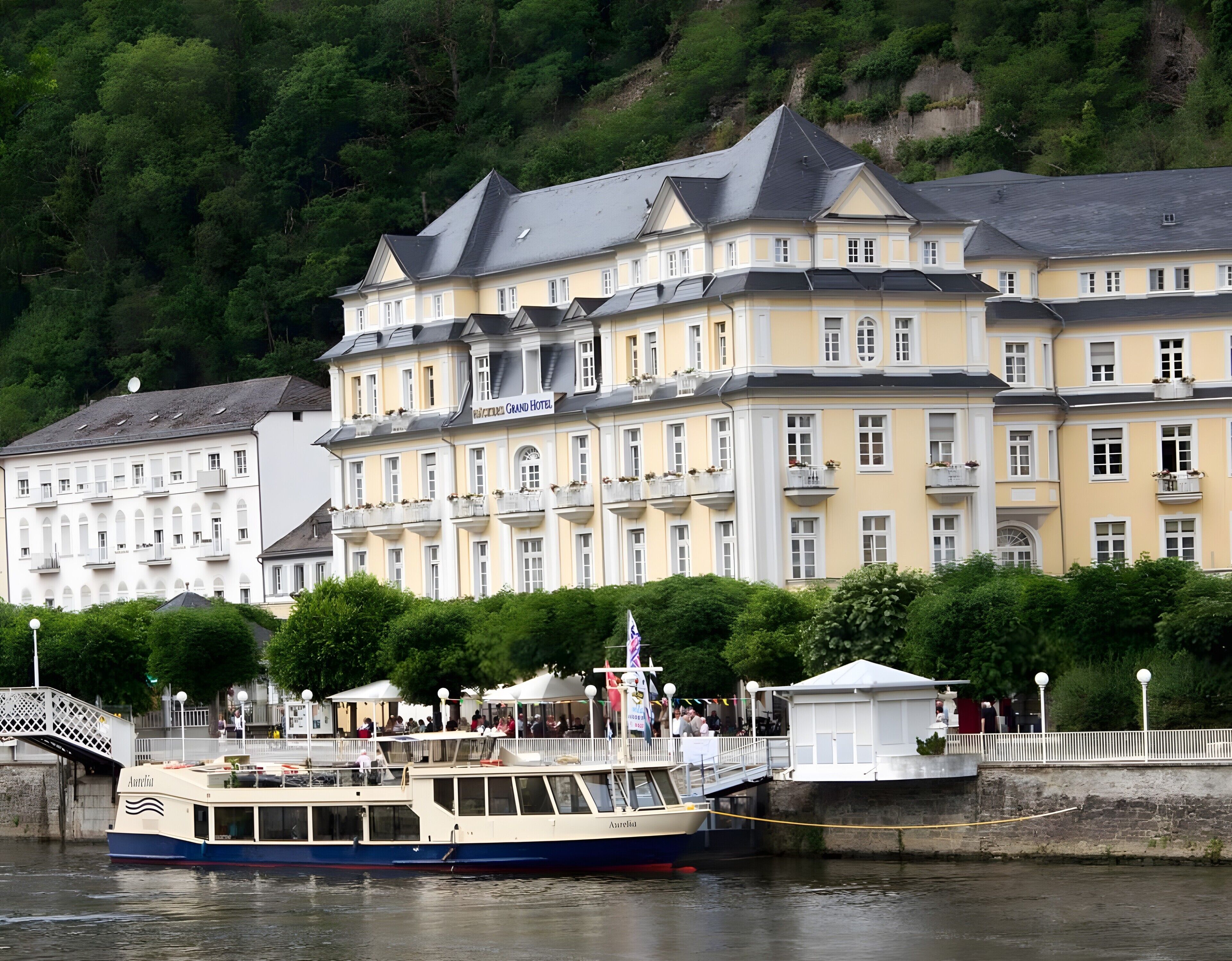A large, multi-story hotel with balconies, a river in front, and a bridge nearby.