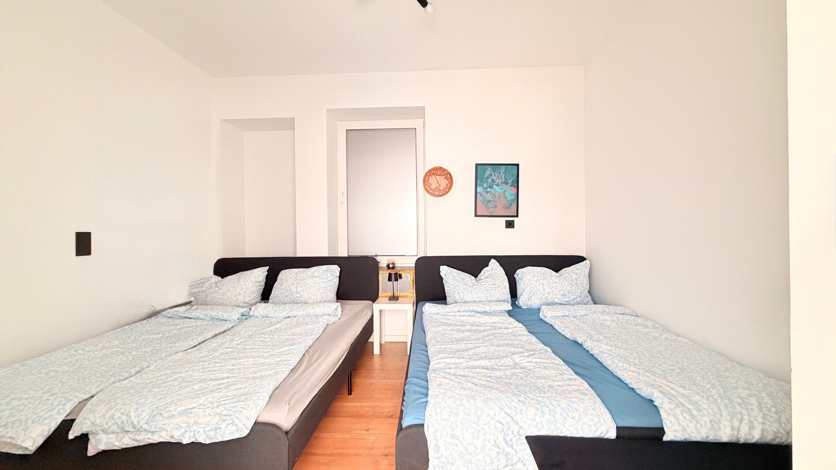 Une chambre avec deux lits simples, une petite table, une horloge et un tableau encadré au mur.