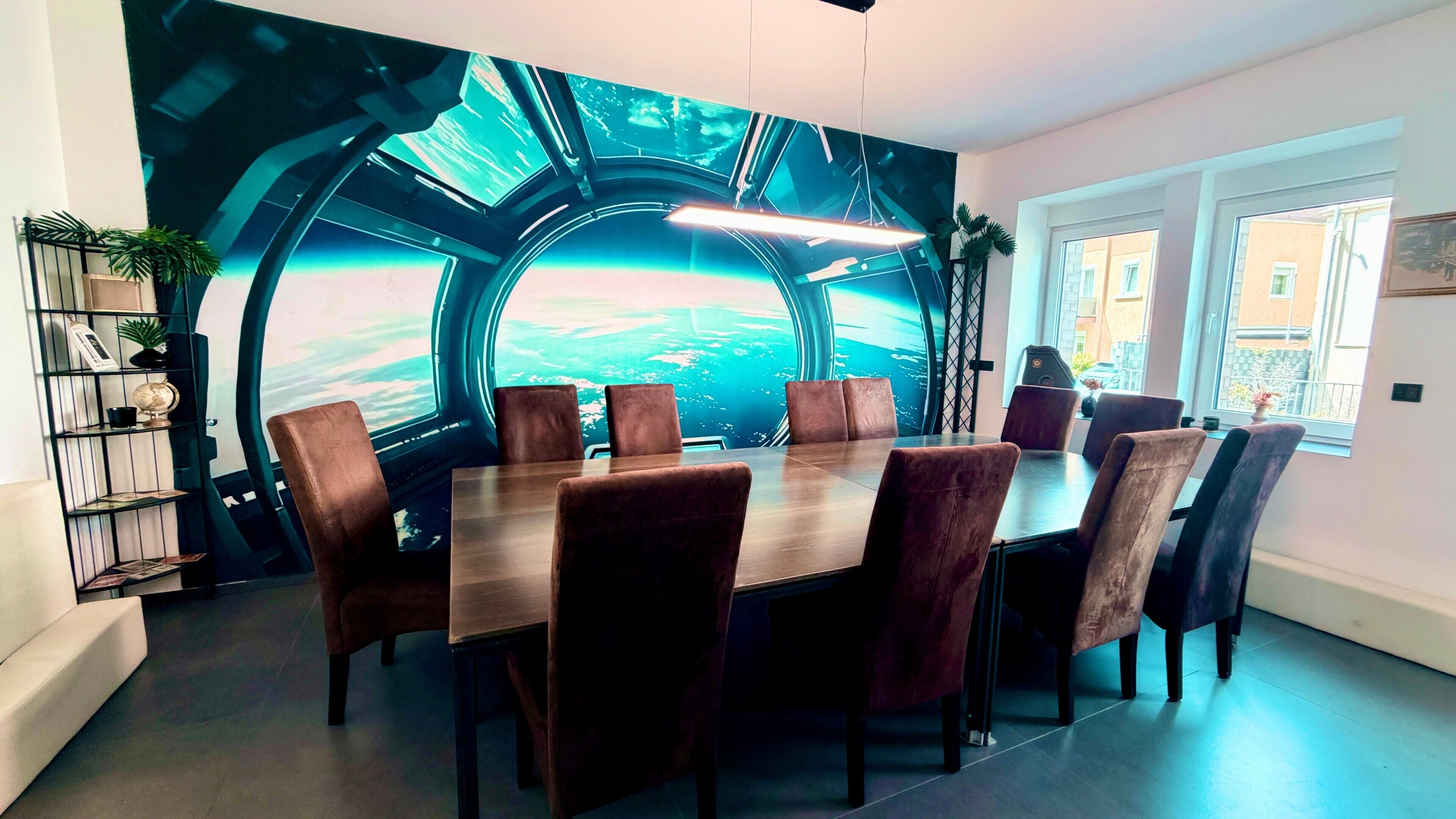 Spisestue med et stort bord, flere stole, et mural af et futuristisk rumskib og udsigt over byen gennem et vindue.
