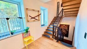 Eine Treppe mit Holzhinweisen und schwarzen Geländern, ein Fenster mit Blick auf Bäume und ein gelber Tisch mit einem Blumenstrauß.