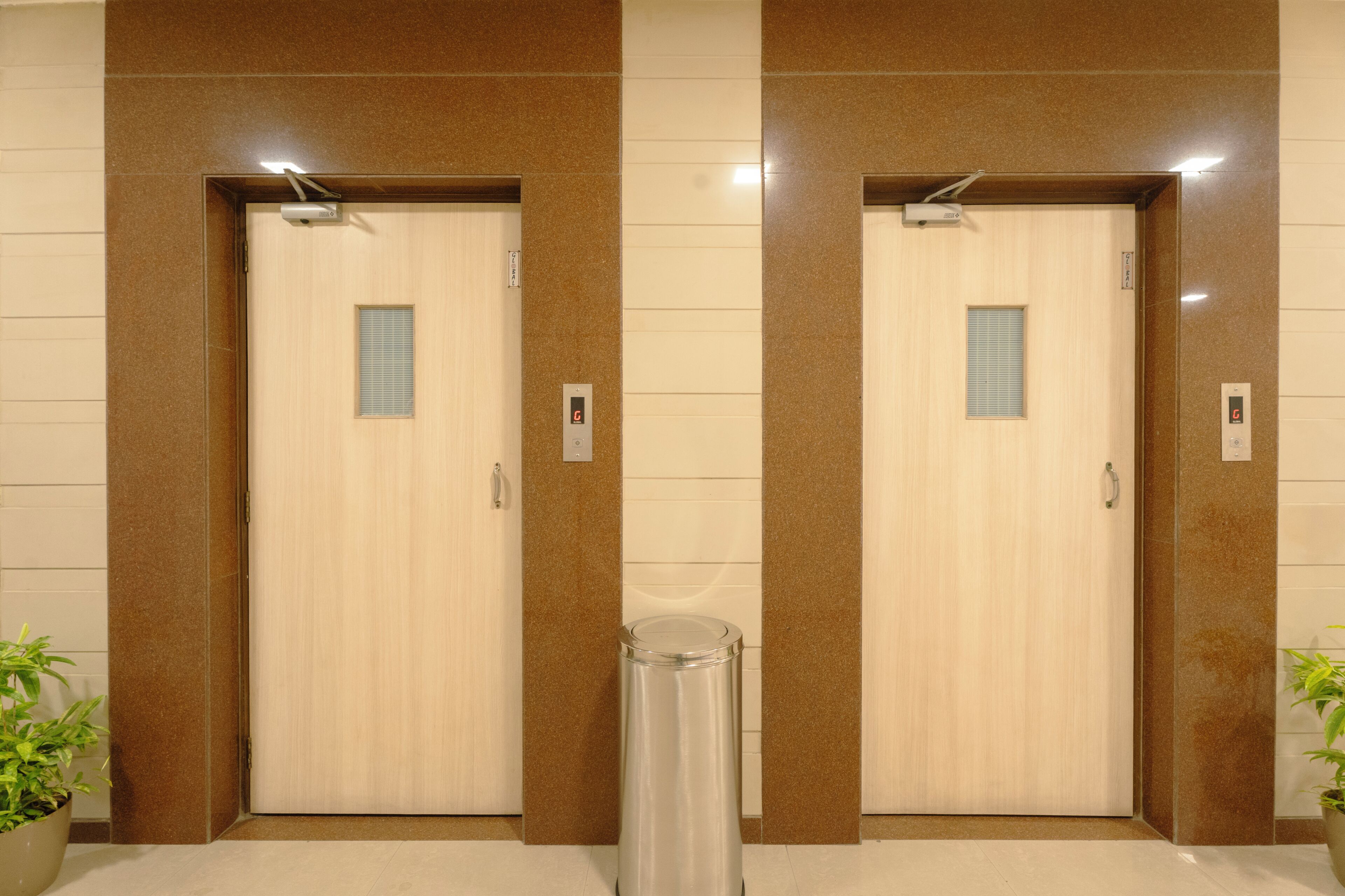 Duas portas de elevador com um aparelho de iluminação acima de cada uma, uma lixeira no centro e uma planta em vaso à esquerda.
