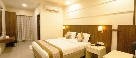 A hotel room with a bed, bedside table, desk, chair, and an air conditioning unit.
