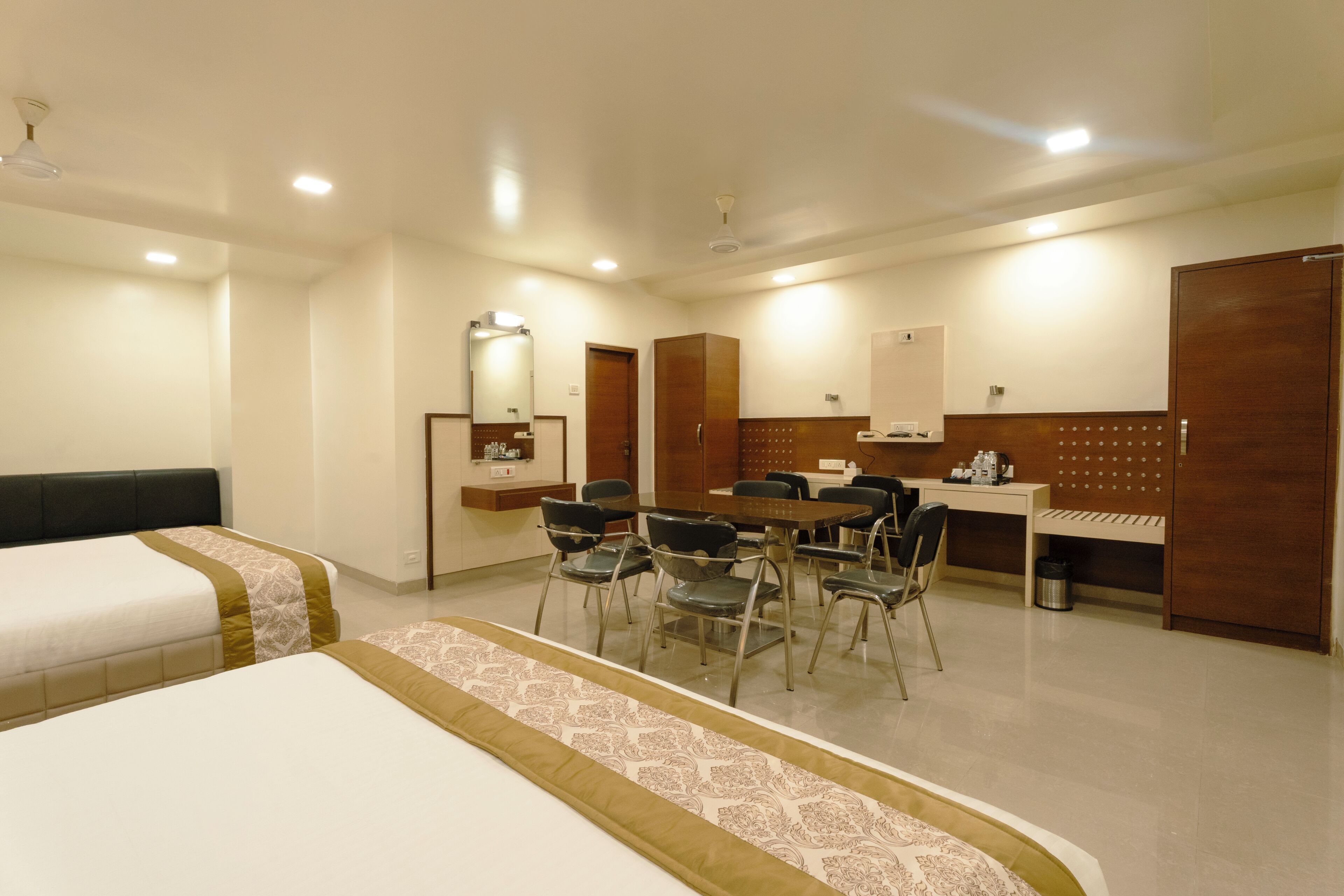 A hotel room with two beds, a dining table with chairs, a small kitchen area, and a mirror.
