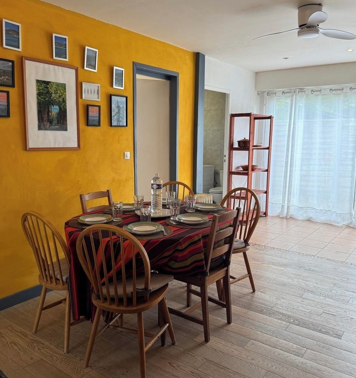 Un coin repas avec une table dressée pour quatre personnes, des chaises en bois, un ventilateur de plafond et une fenêtre avec des rideaux.