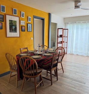 Un comedor con una mesa dispuesta para cuatro, sillas de madera, un ventilador de techo y una ventana con cortinas.