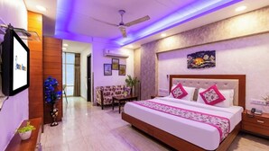 A hotel room with a large bed, a desk, a chair, a ceiling fan, and a TV. - Oly Emporio Near NDLS (New Delhi)