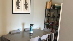 A dining area with a glass table, white chairs, a teal vase, and a bookshelf with books and decorative items. - Hackney Road Studio Near Shoreditch Markets (London)