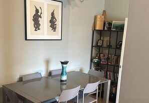 Ein Essbereich mit einem Glastisch, weißen Stühlen, einer türkisblauen Vase und einem Bücherregal mit Büchern und Dekoration.