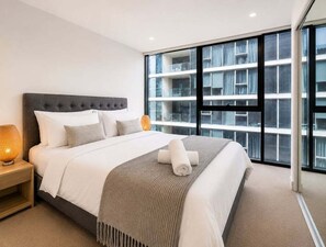 A modern hotel room with a large bed, a nightstand, a lamp, and a view of the building's exterior through large windows. - High-Rise Retreat Near South Bank (Brisbane)