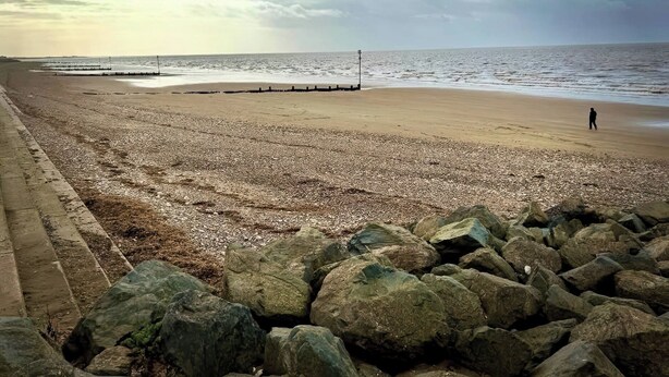 Une plage de galets avec un premier plan rocheux, une personne qui marche et une mer calme sous un ciel partiellement nuageux.
