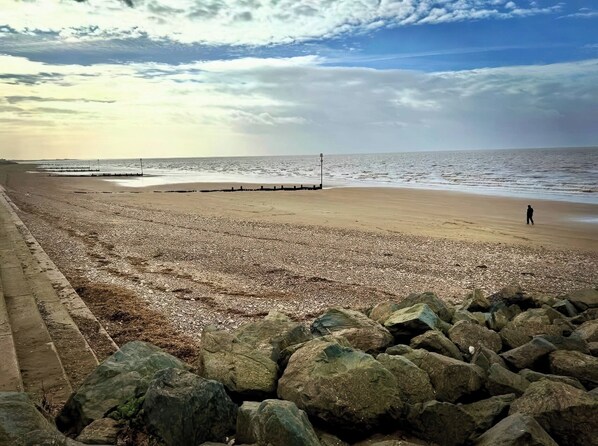 A pebbled beach with a rocky foreground, a person walking, and a calm sea under a partly cloudy sky. - 3b Hideaways (Hunstanton)