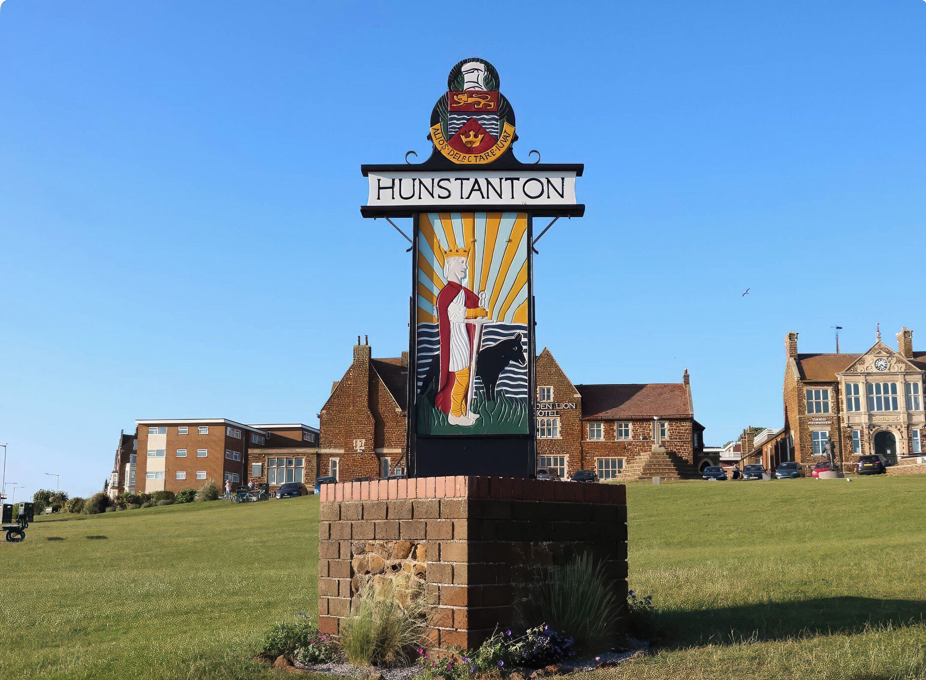 Un panneau indicateur portant l’inscription « HUNSTANTON » et affichant des armoiries, placé devant un bâtiment de style architectural traditionnel.