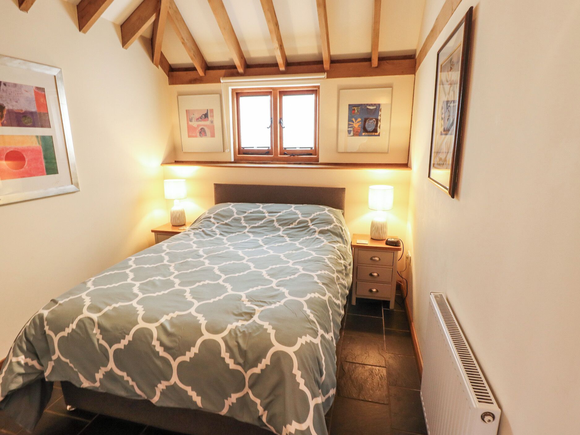 Un dormitorio con una cama, dos mesitas de noche con lámparas, una ventana y un radiador.