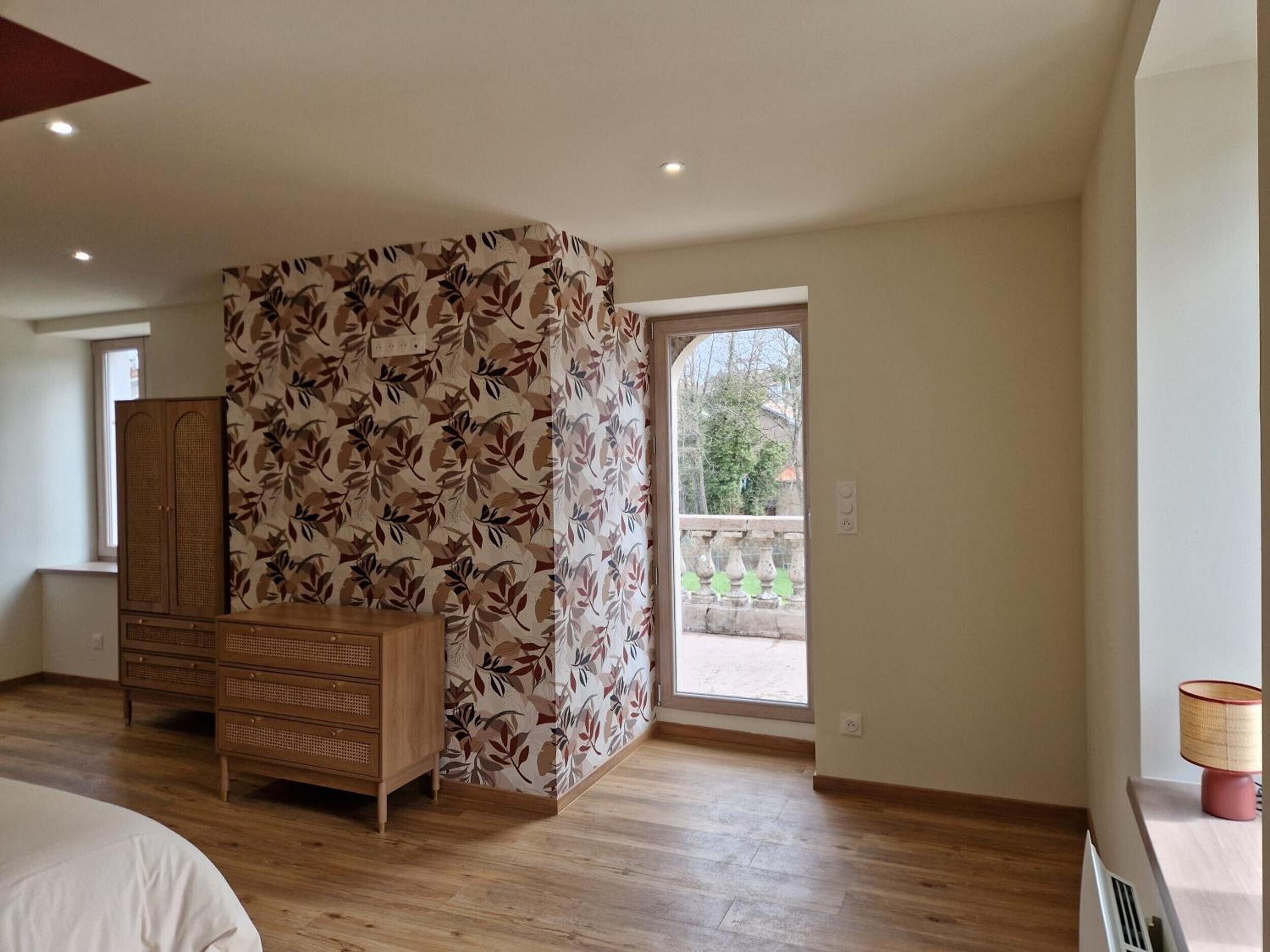 A bedroom with a wooden wardrobe, a bed, a window with a view of a balcony, and a door leading outside.
