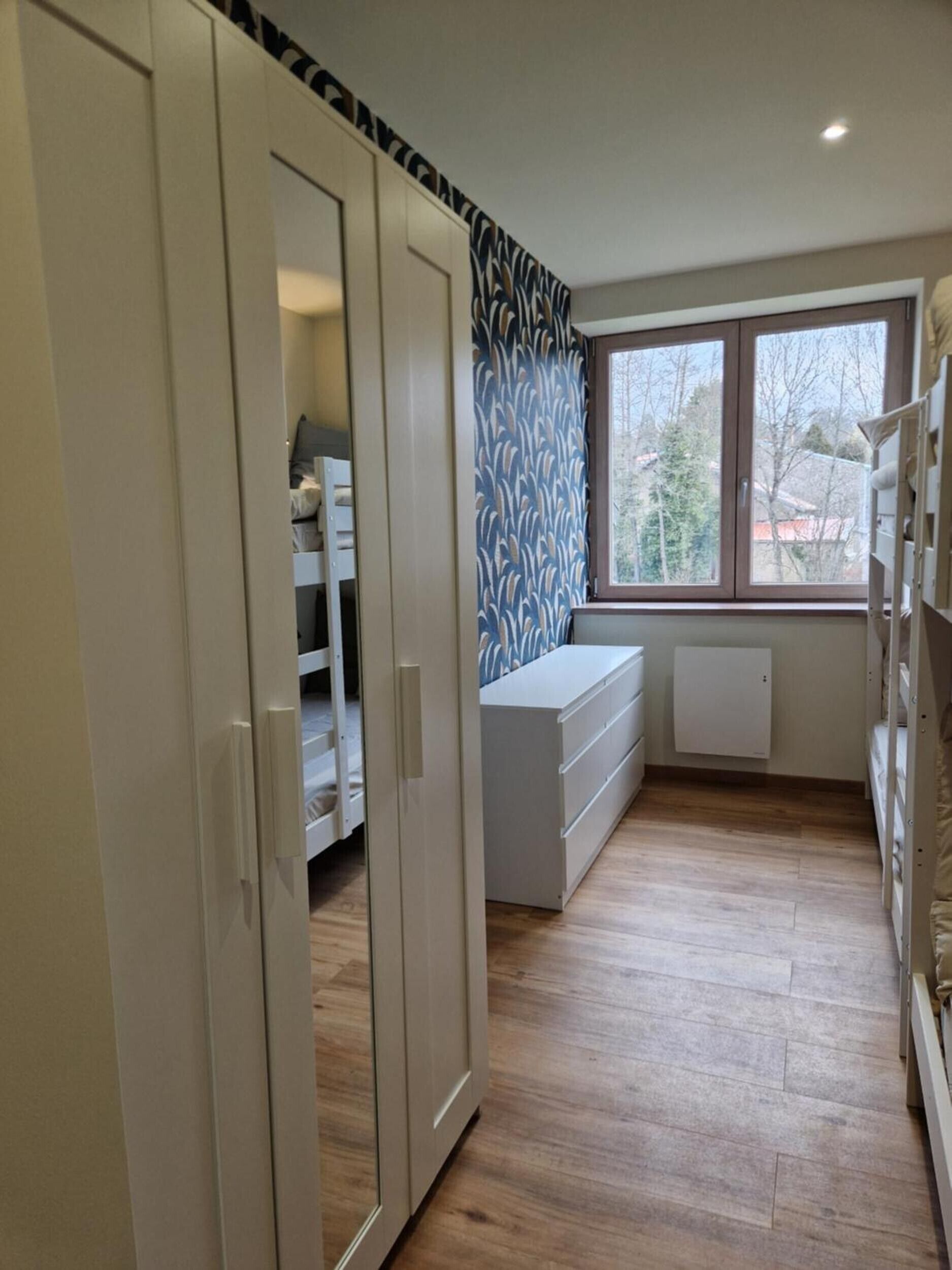 Een kamer met een kledingkast, een witte dressoir, een radiator en een raam met uitzicht op bomen.