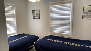Un dormitorio con dos camas, paredes blancas, ventilador de techo y una cita inspiradora enmarcada.