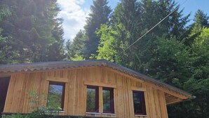 Uma cabana de madeira com telhado inclinado, rodeada por folhagem densa e árvores altas.