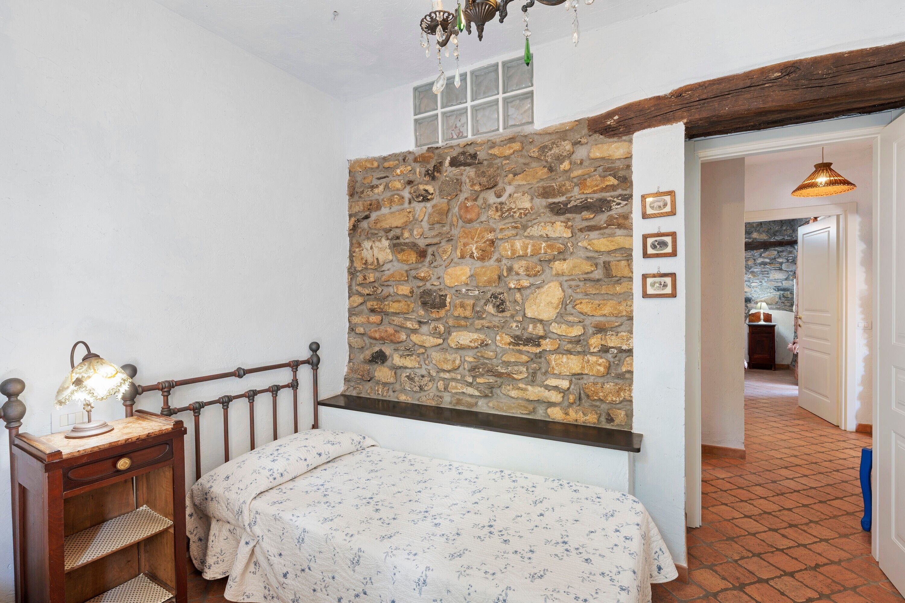 A bedroom with a stone wall, a bed with a floral bedspread, a nightstand with a lamp, and a wooden ceiling beam.