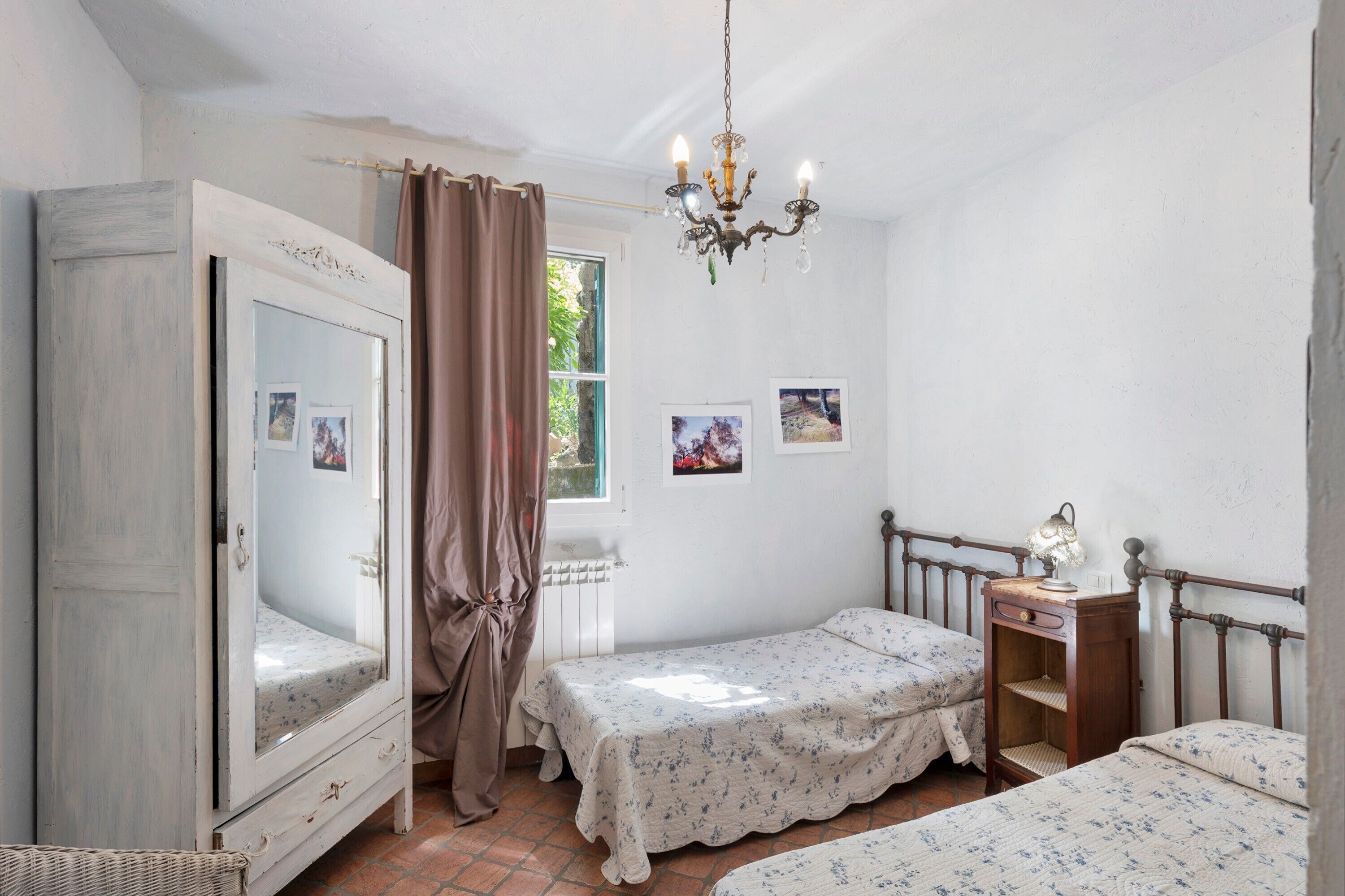 A bedroom with two beds, a dresser, a mirror, a chandelier, and a window with curtains.