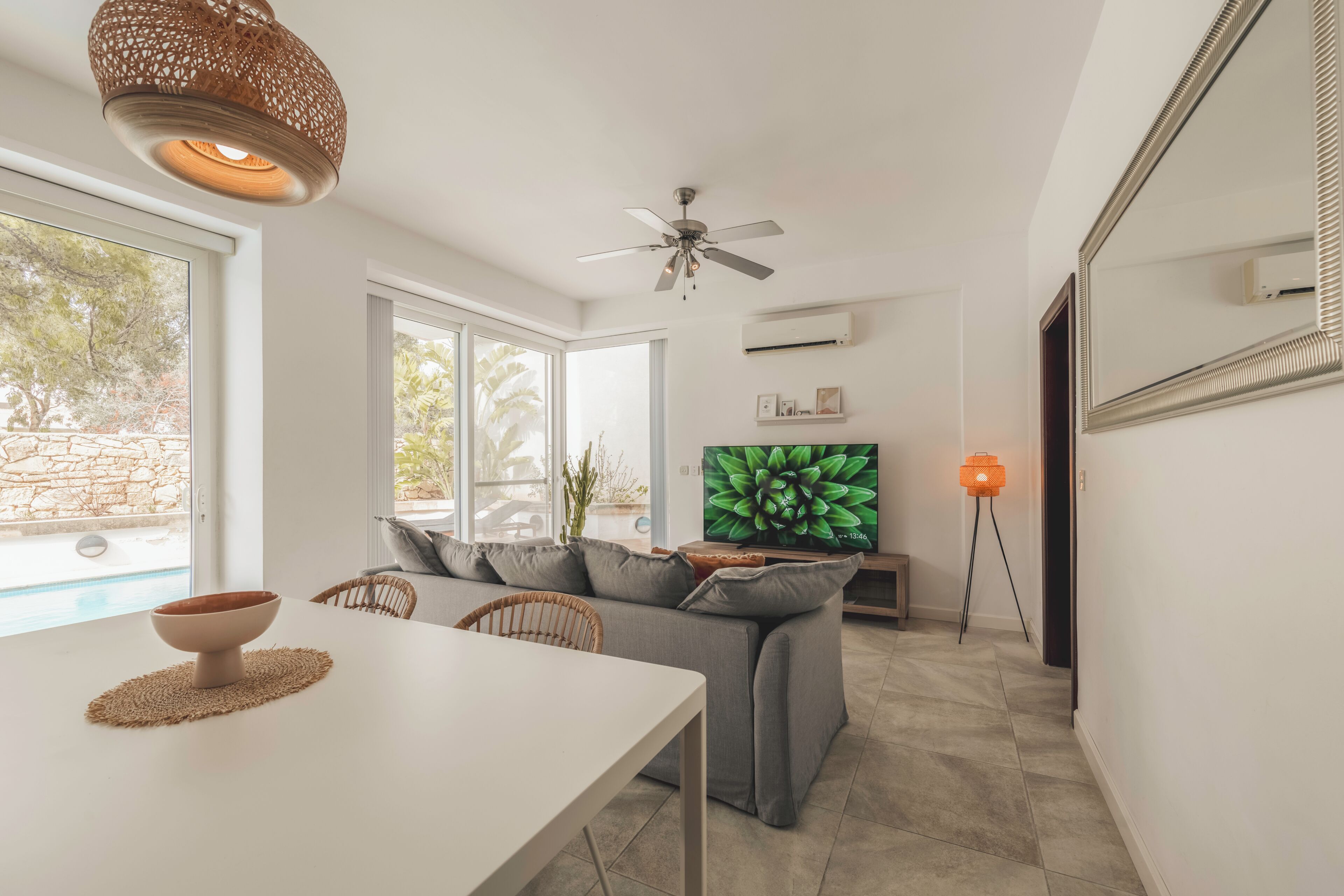 A modern living room with a dining table, grey sofa, ceiling fan, TV, and a view of the outdoors.