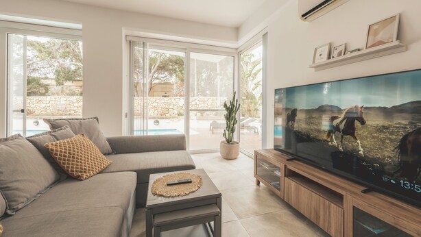 A modern living room with a sofa, TV, and a view of a pool and outdoor area. - Villa Marni (Munxar)
