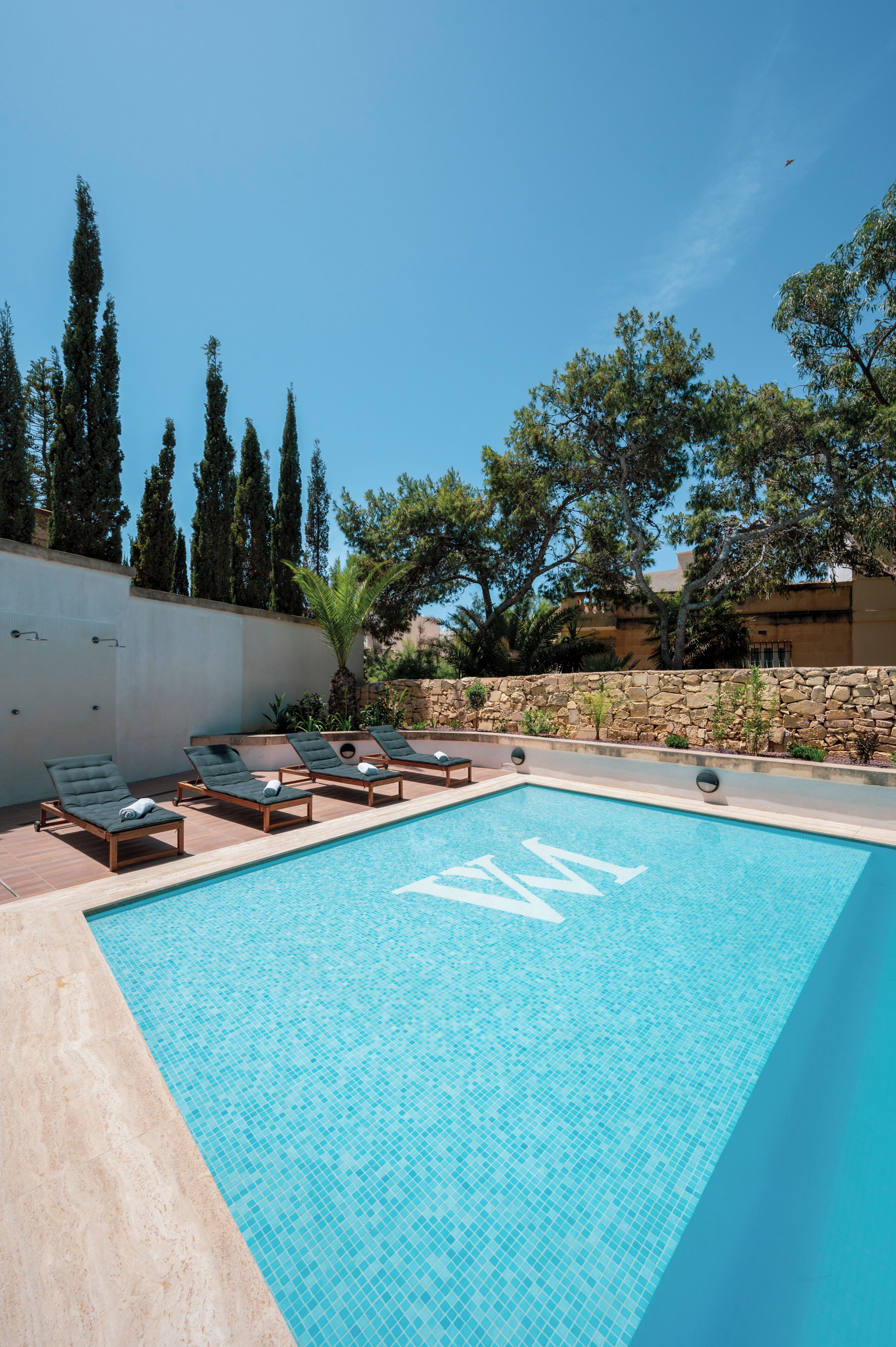 Una piscina con lettini, un muro in pietra e alberi sullo sfondo.
