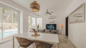 A modern living room with a dining table, wicker chairs, a ceiling fan, a TV, and a large window overlooking a pool. - Villa Marni (Munxar)