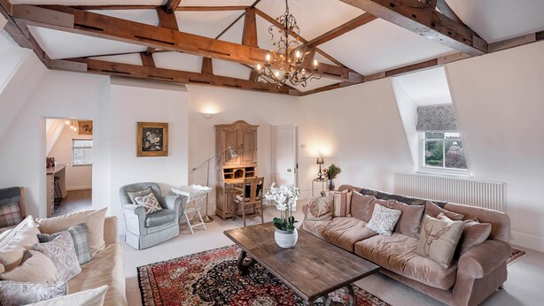 Ein geräumiger Loft mit freiliegenden Holzbalken, einem großen Fenster, einer gemütlichen Sitzecke mit Sofa und Sesseln sowie einem dekorativen Kronleuchter.